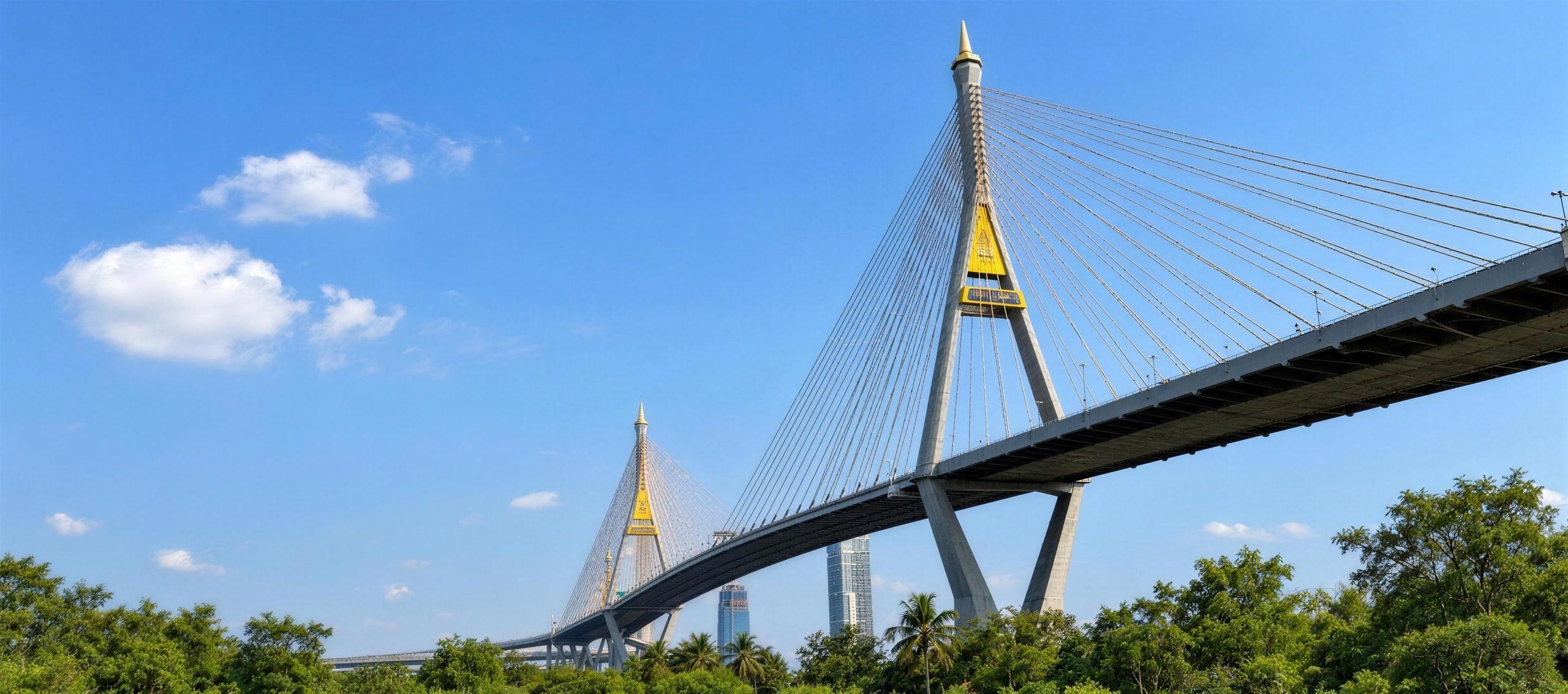 The Bhumibol Bridge, a cable-stayed bridge with gold-tipped pylons, rising over trees in Bangkok under a clear blue sky